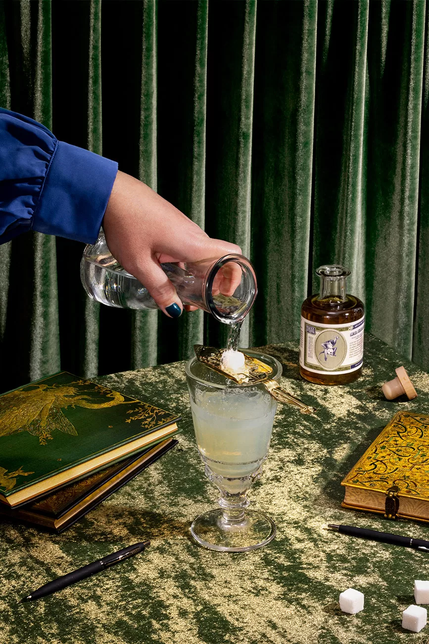 A woman's hand pours water over a sugar cube into a vintage glass with absinthe. Narrative food photography with journals and pens and the product of St George Spirits bottle. Green fairy.