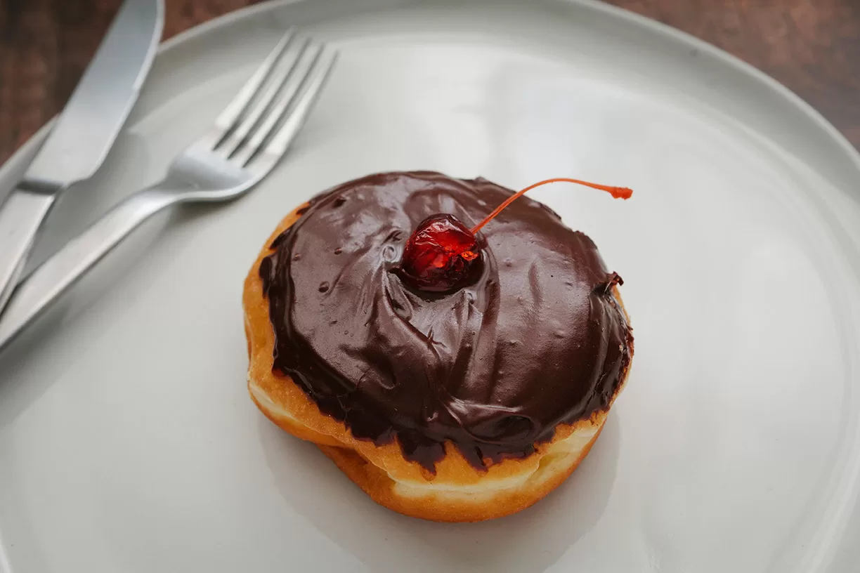 Boston Creme donut topped with a bright red cherry on a white plate with a knife and fork