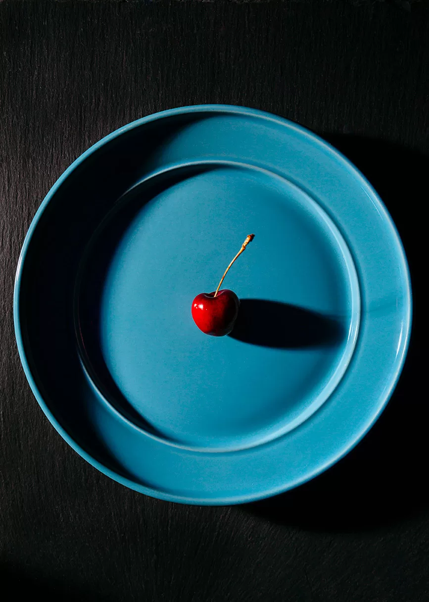 Single bright red cherry with stem on a robin blue plate with dark dramatic shadows and lighting. Milwaukee food photographer