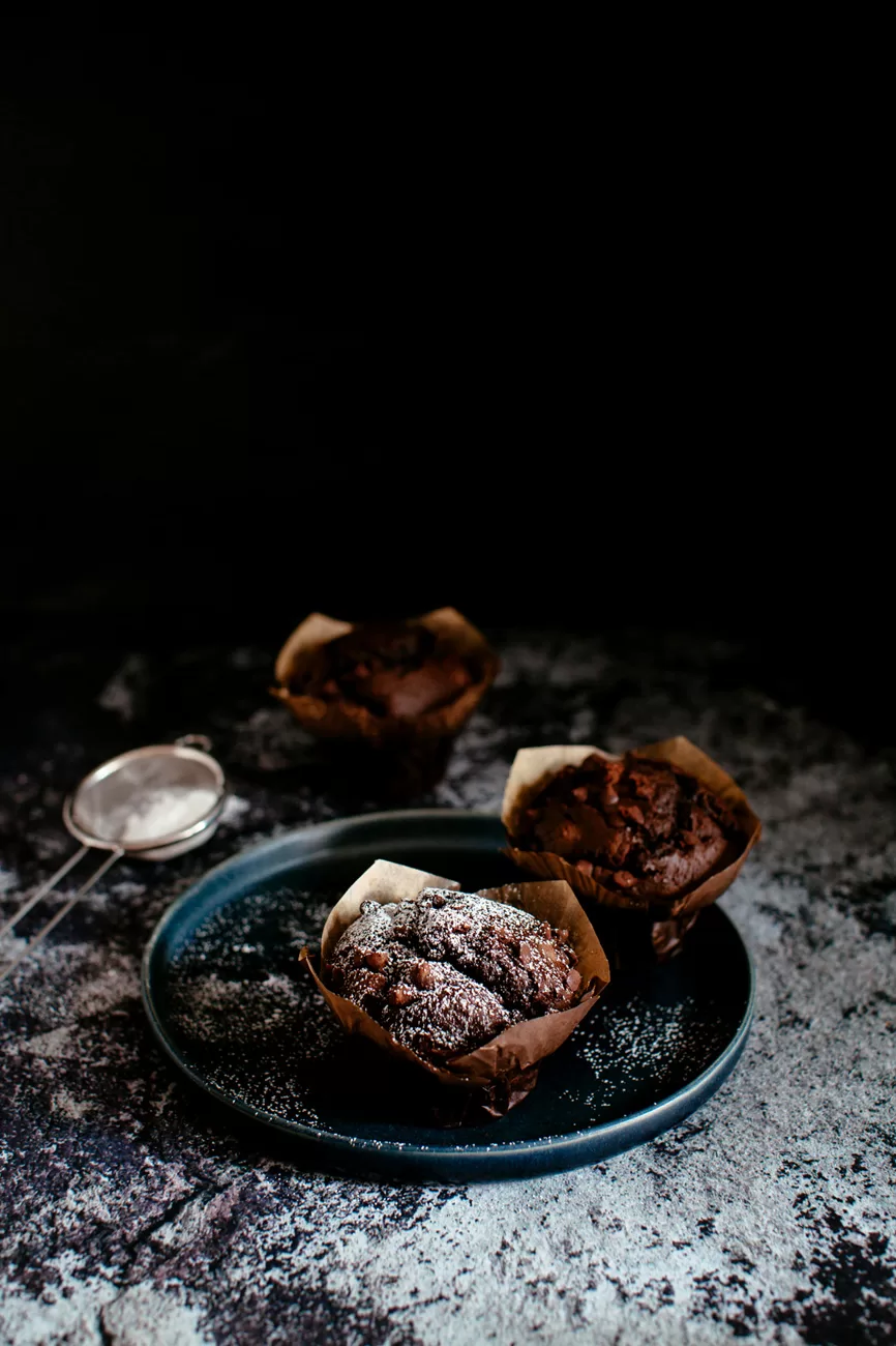 Double chocolate chip muffins dusted with powdered sugar set on a cobalt blue plate and concrete tabletop.
