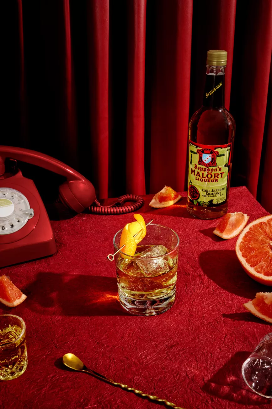 Jeppson’s Malört bottle product photography in a narrative scene with a boulevardier cocktail topped with a curled orange peel complimented with grapefruit slices, shot glasses, and a vintage rotary phone. Chicago's Drink.