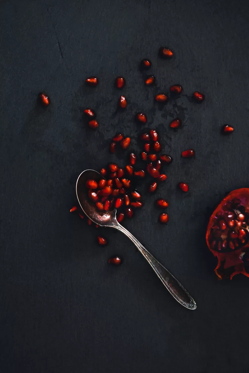 Pomegranate Seeds spread out with a vintage silver spoon and half of the fruit on a black slate board. Dark natural lighting