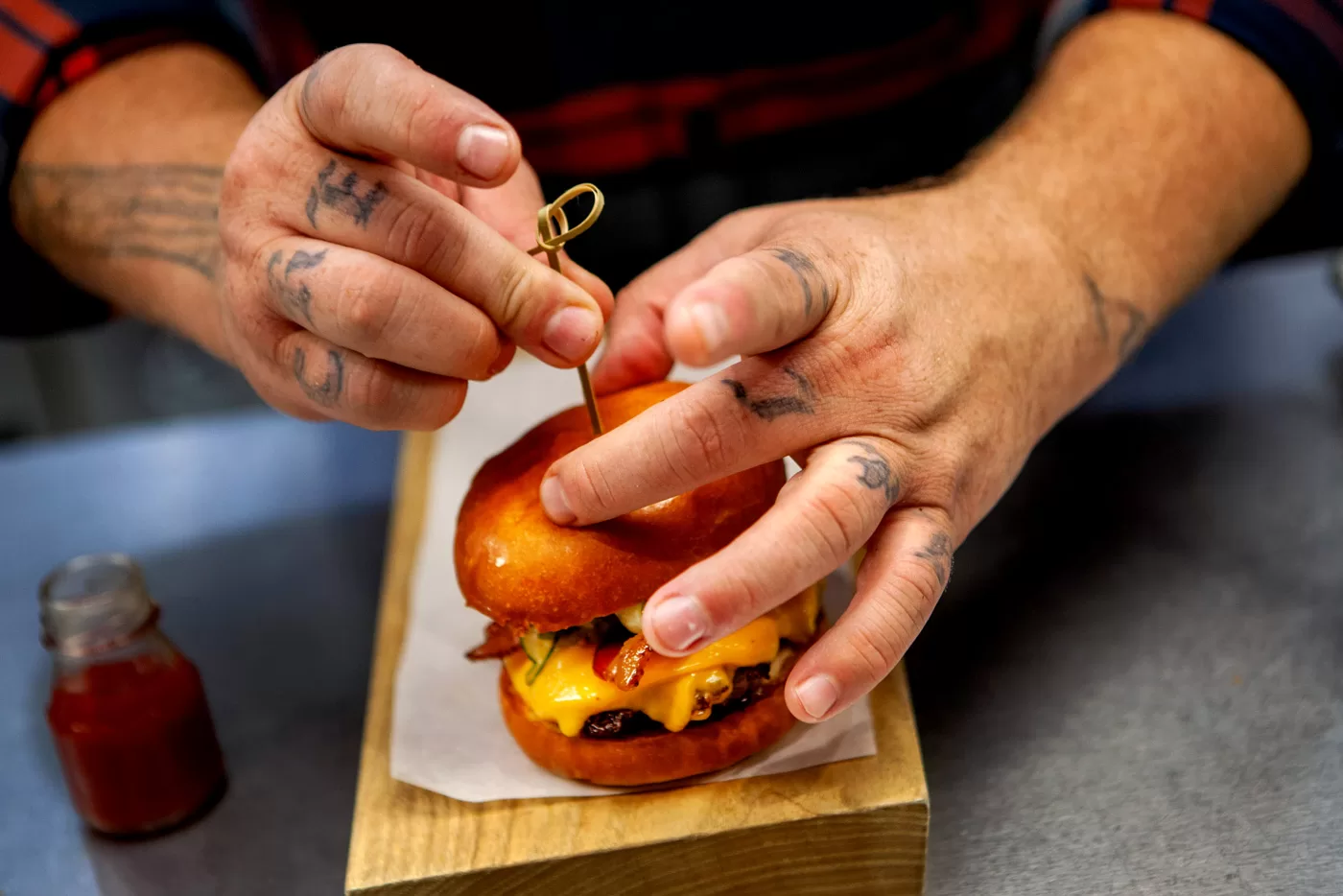 Restaurant Chef with Knuckle tattoos preparing a cheeseburger on a wooden tray slab with a side of mini ketchup